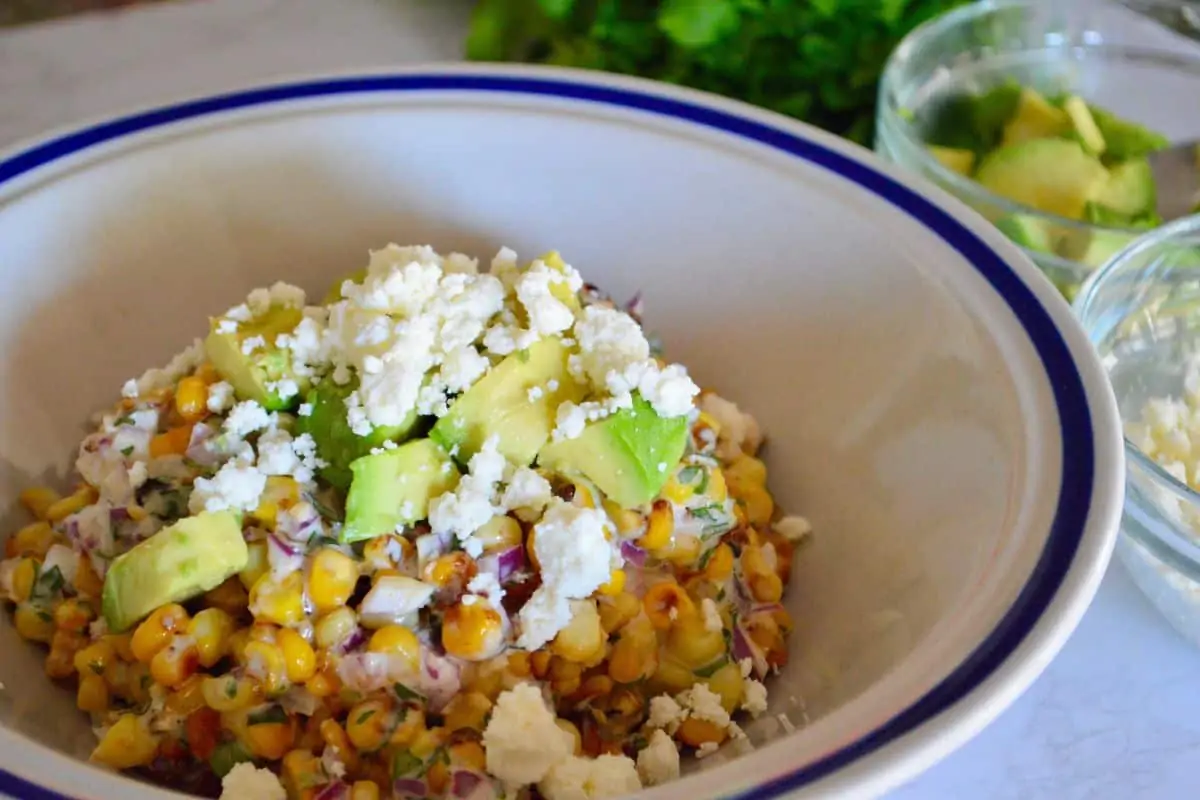 Mexican street corn salad with avocado in a white bowl with avocado and cotija cheese on top. 