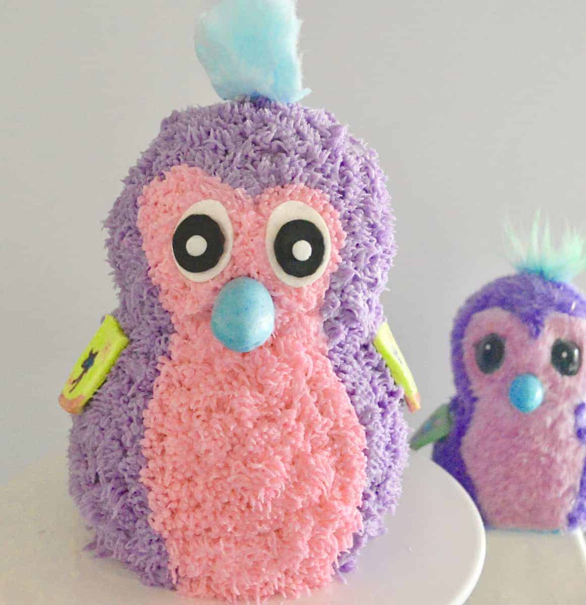 Purple and pink Hatchimals cake on a white pedestal with hatchimal toy in the background. 