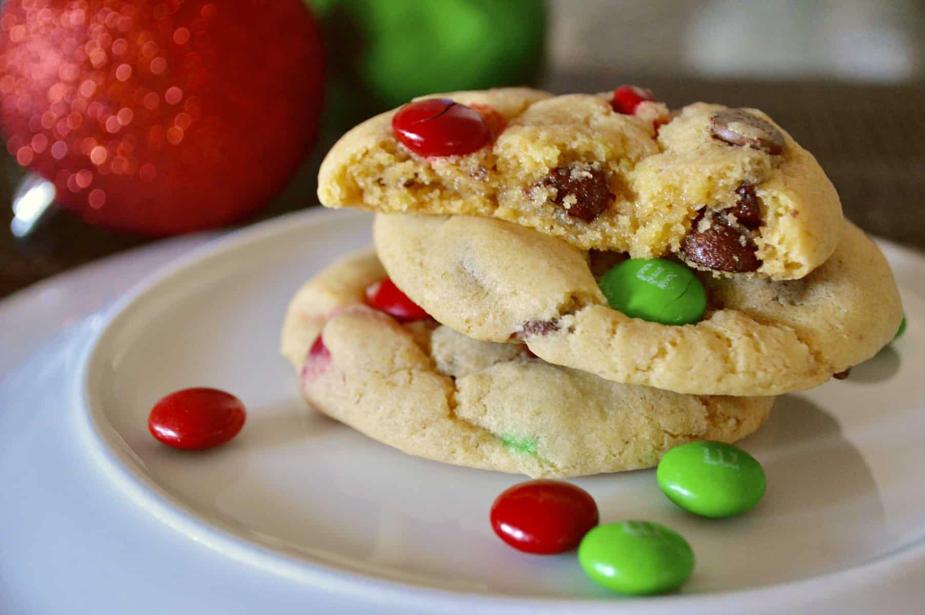 Christmas M&M Cookies on a white plate with red and green ornaments in the background 