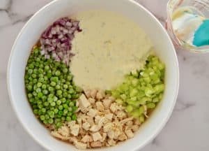 greek yogurt dressing in a white bowl along with pasta, chicken, peas, and red onion.