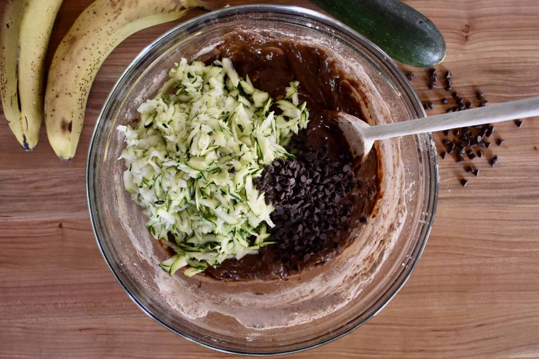overhead photo of grated zucchini and chocolate chips in the bread batter in a glass bowl with a wooden spoon. 