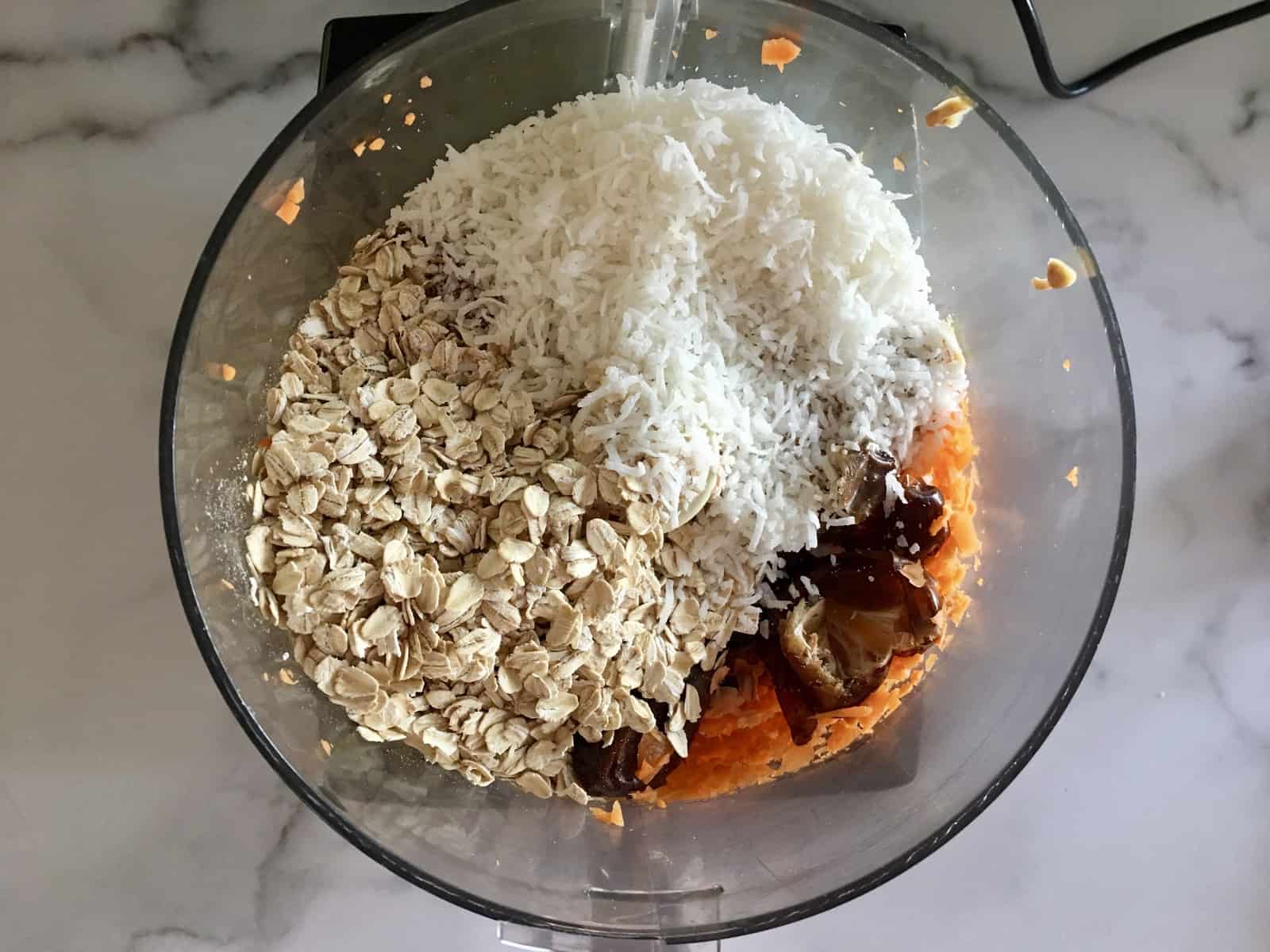 overhead photo of food processor with oats, dates, shredded coconut, carrot, and peanut butter