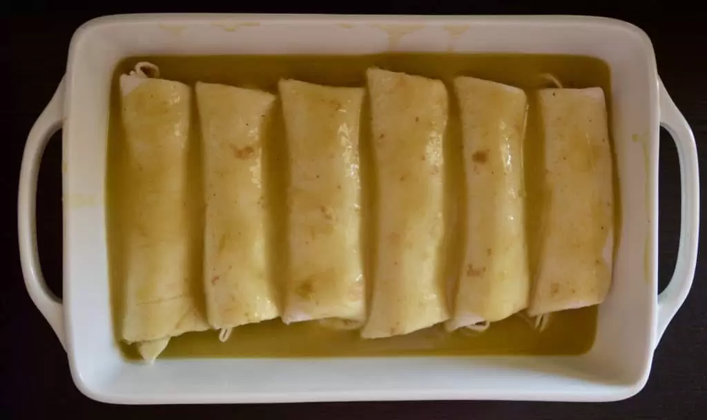 green chili enchilada sauce poured over top of six burritos in a white baking dish. 