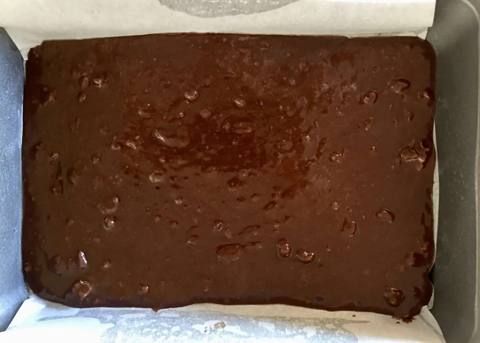 Brownie batter spread along the bottom of a baking pan lined with parchment paper. 