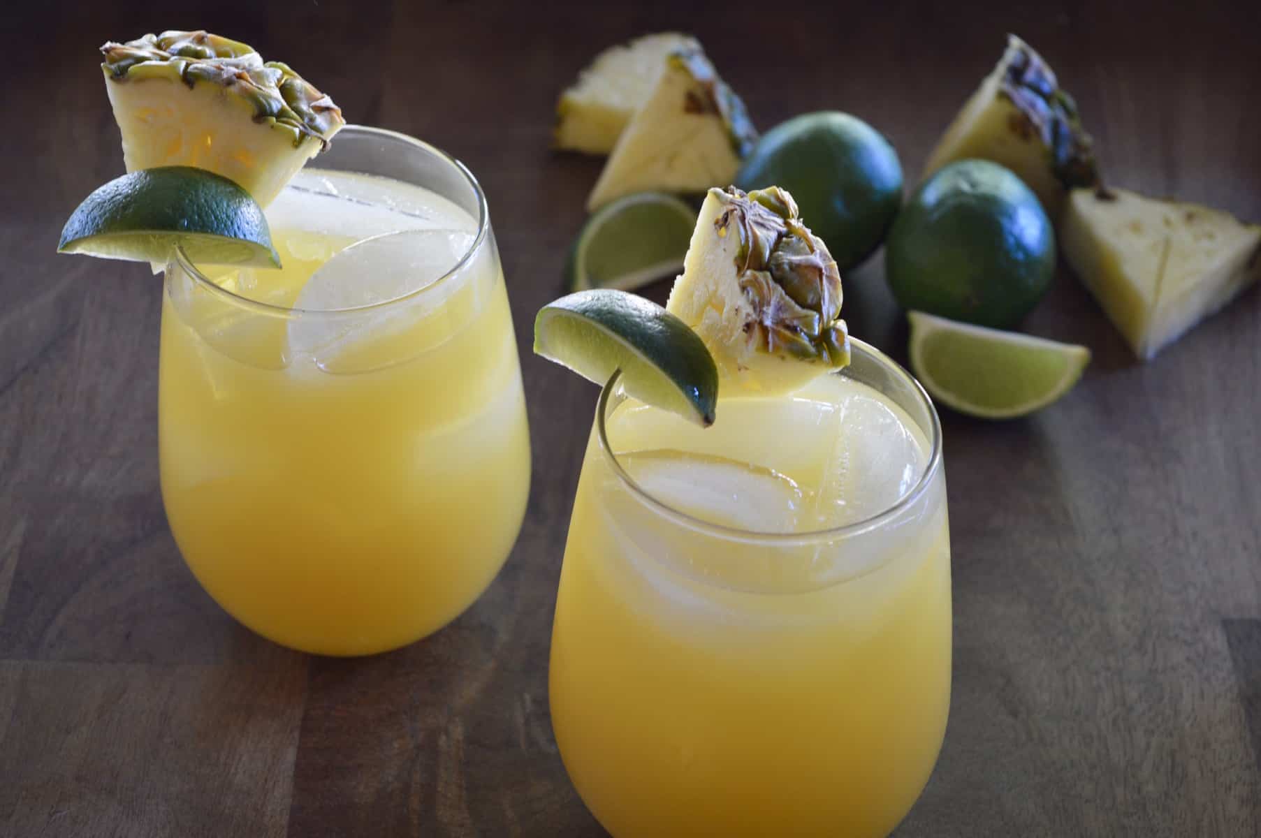 Pineapple margaritas on a wood platter with pineapples slices and lime wedges. 