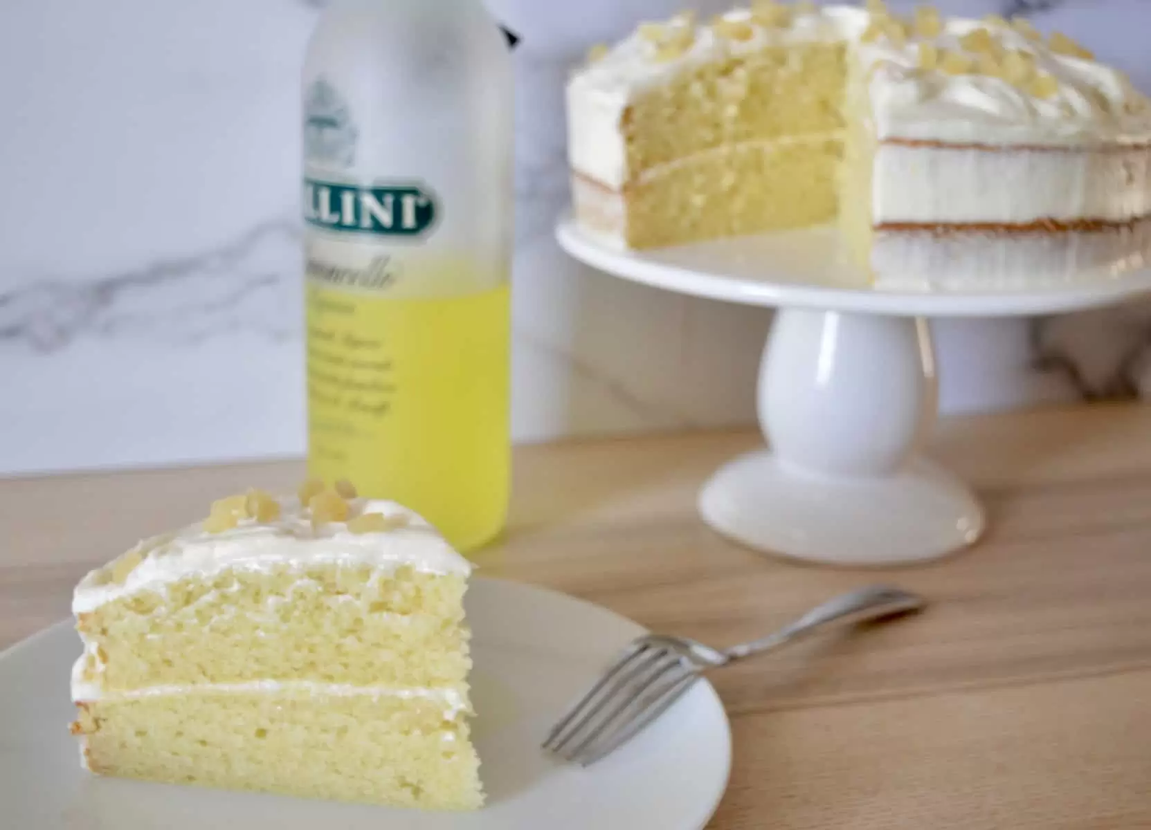 Limoncello Ricotta Cake on a pedestal with a bottle of Limoncello liqueur in the background. 