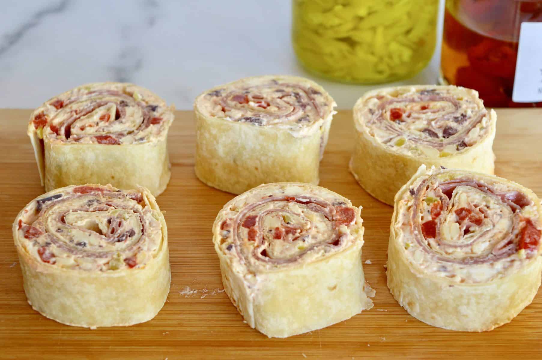 pinwheels on a cutting board with pepperoncini and roasted bell peppers in the background.