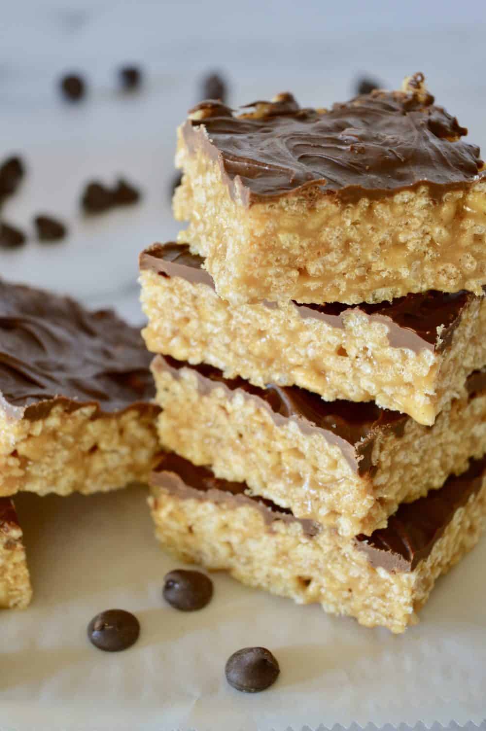 Caramel Peanut Butter Rice Krispie Treats stacked up high on a white top with chocolate chips in the background. 