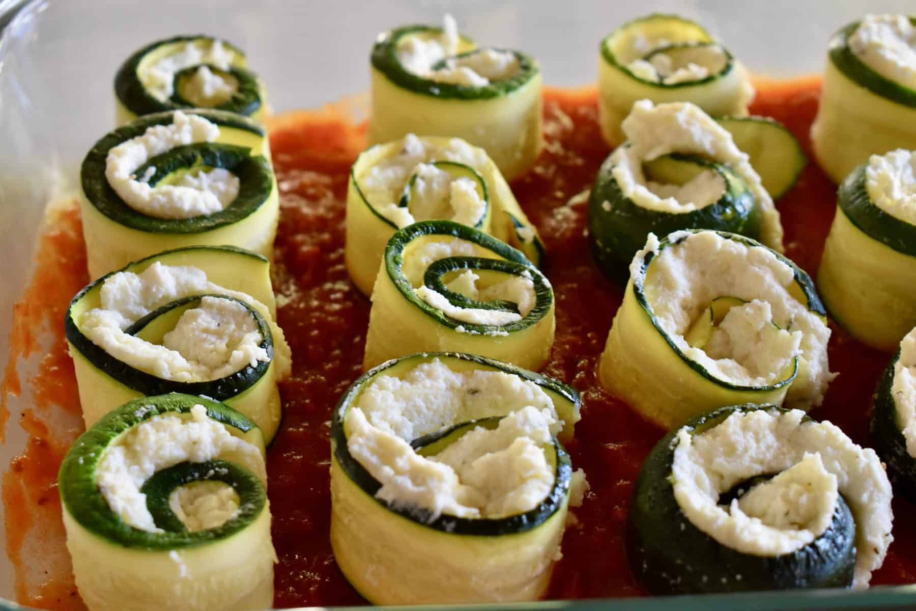 Zucchini ricotta rolls up standing upright in a glass baking pan with marinara sauce on the bottom. 