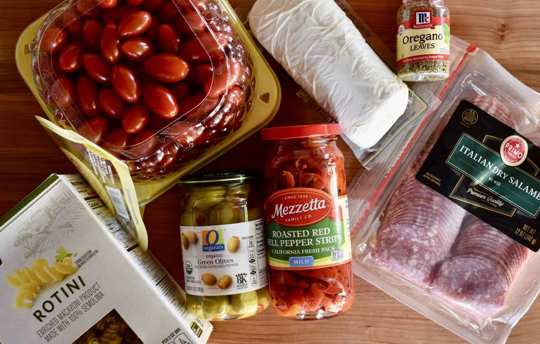 Overhead photo of wood cutting board with rotini, salami, tomatoes, mozzarella, green olives, roasted red peppers, and oregano.  