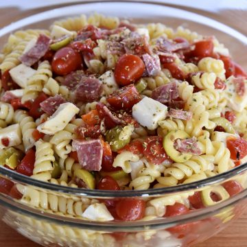 Rotini pasta in a clear bowl with toppings and dressings tossed in it.