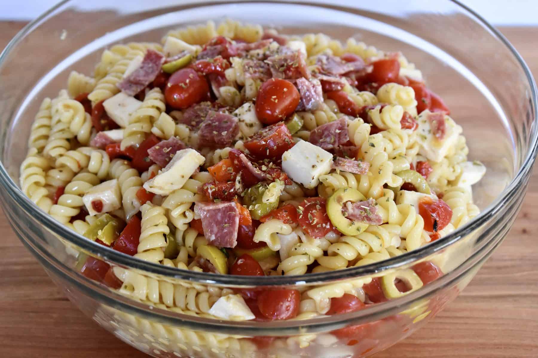 Rotini pasta in a clear bowl with toppings and dressings tossed in it. 