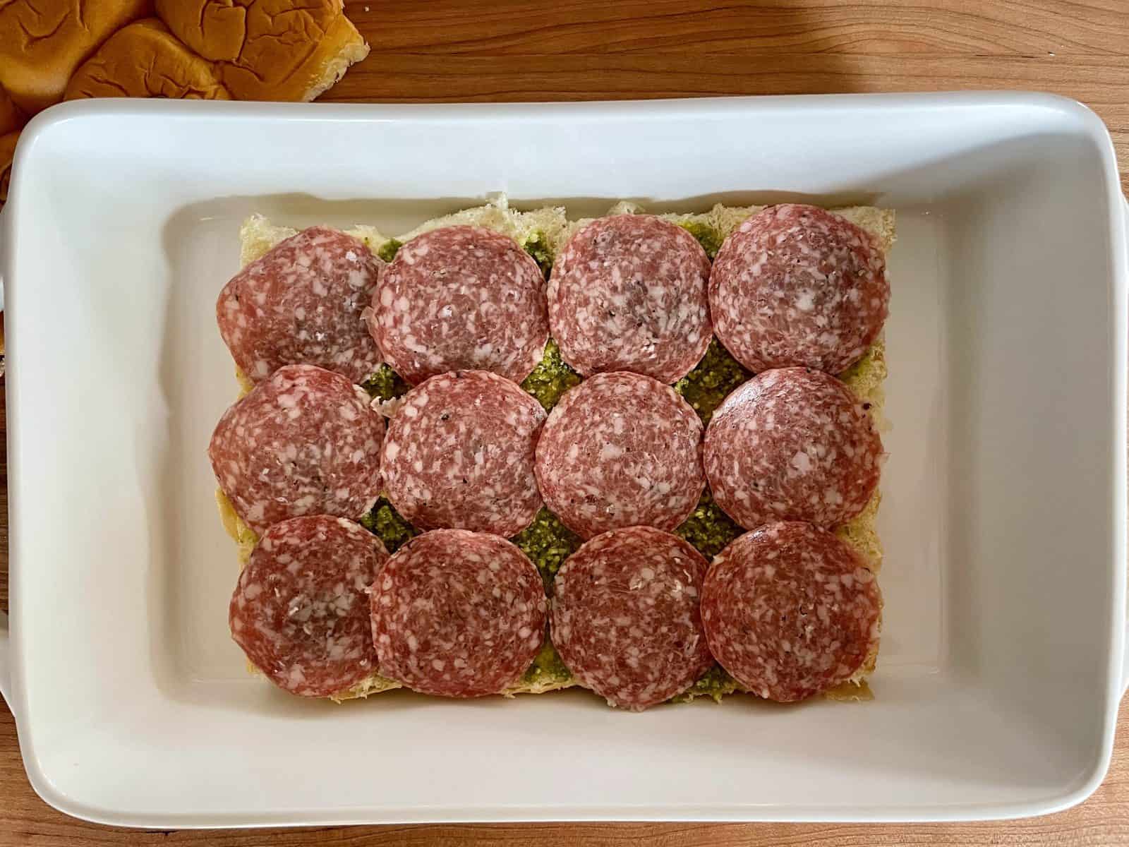 Overhead photo of salami layered over top of the pesto on the bottom half of the rolls. 