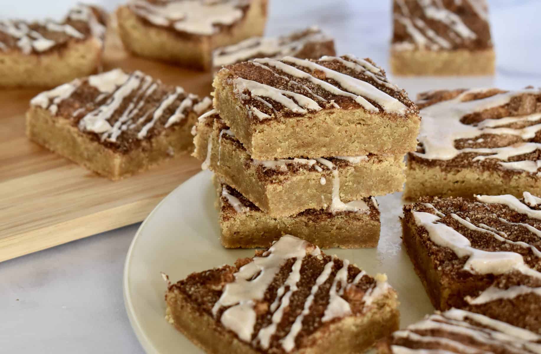 Cinnamon sugar blondies stacked high on a white plate with glaze drizzled over top. 