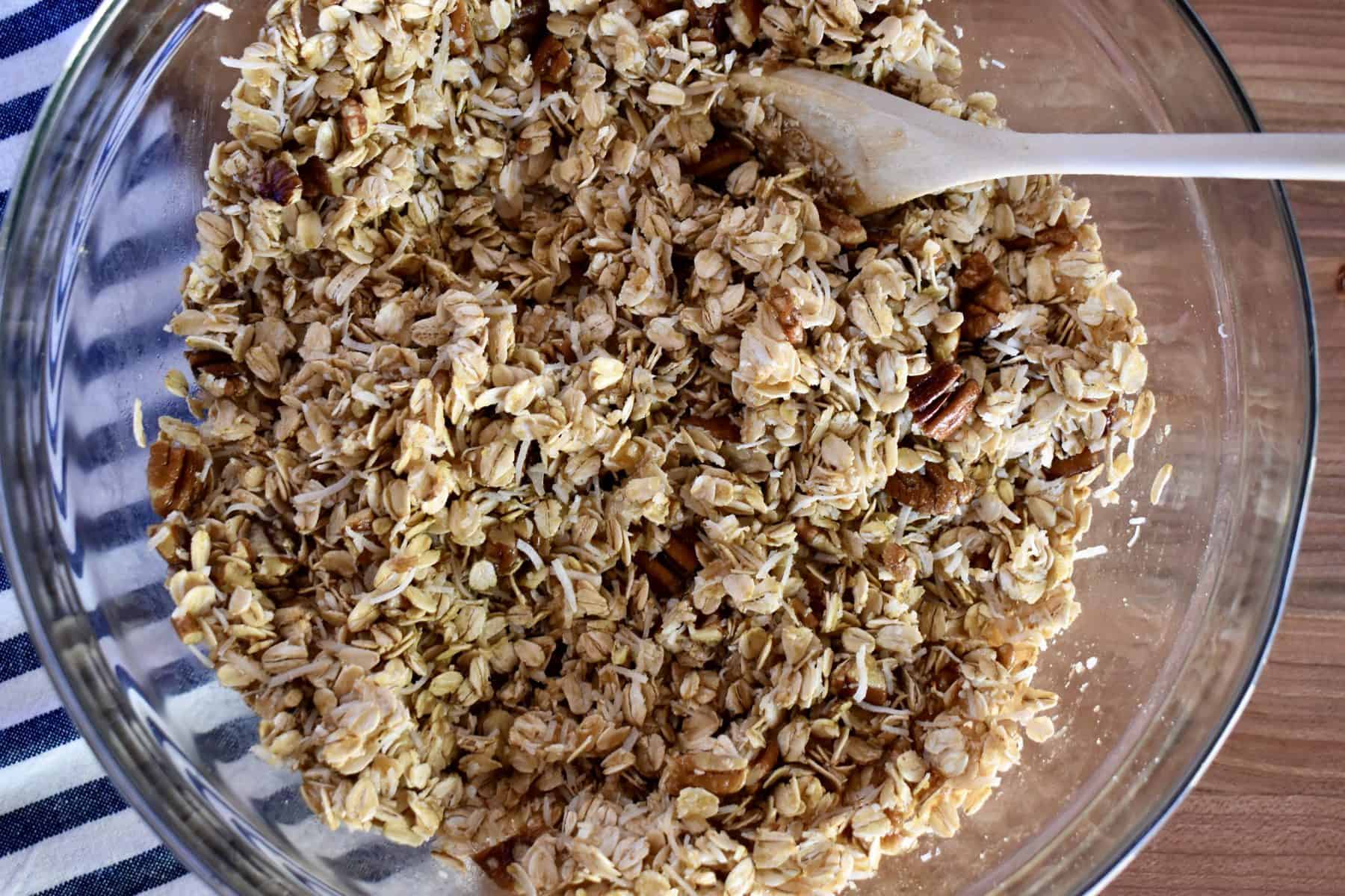overhead photo of coconut pecan granola in a glass bowl with wooden spoon. 