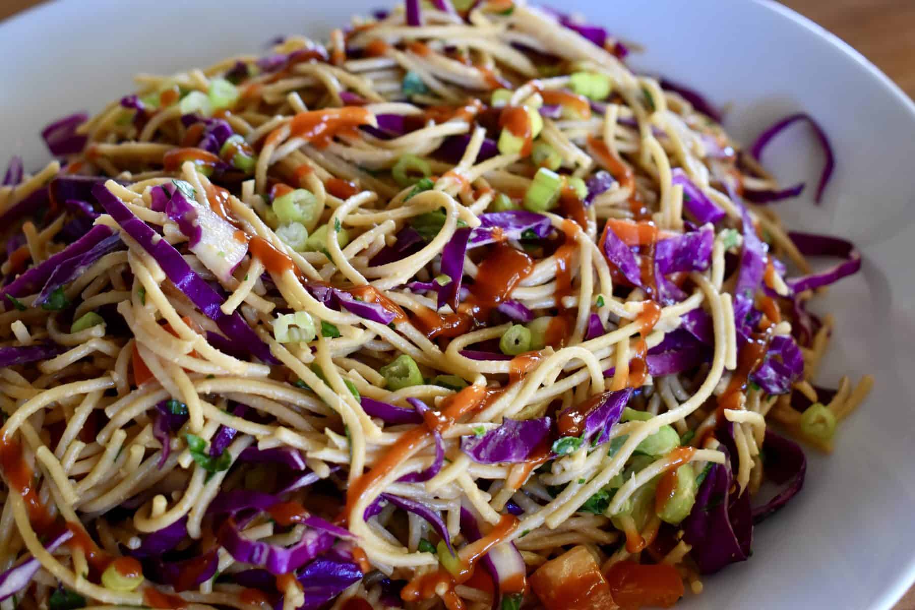 Asian noodle salad in a white bowl with siracha drizzled on top. 