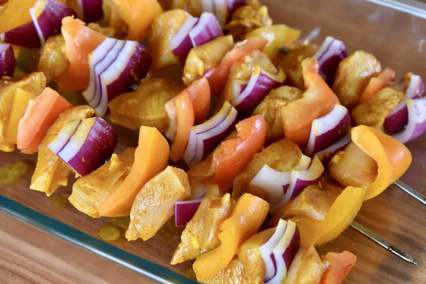 marinated chicken, red onion, and bell pepper on metal skewers in a glass container. 