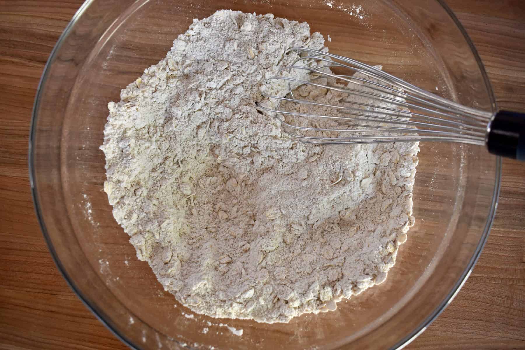 overhead photo of flour, oats, and sugar in a glass bowl with a whisk on a wood cutting board.