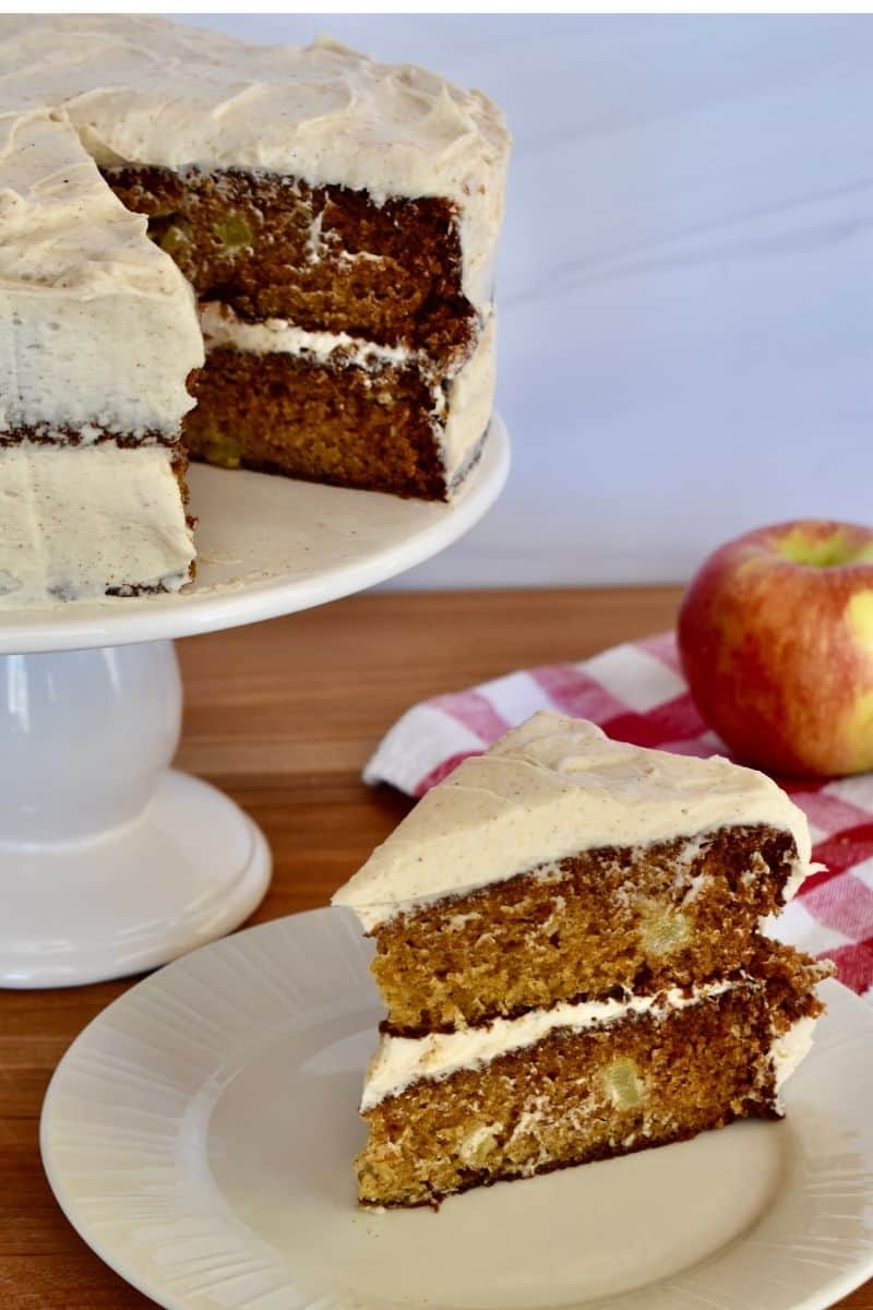 Apple Spice Cake with Cinnamon Buttercream Frosting on a white pedestal with a slice on another white plate.