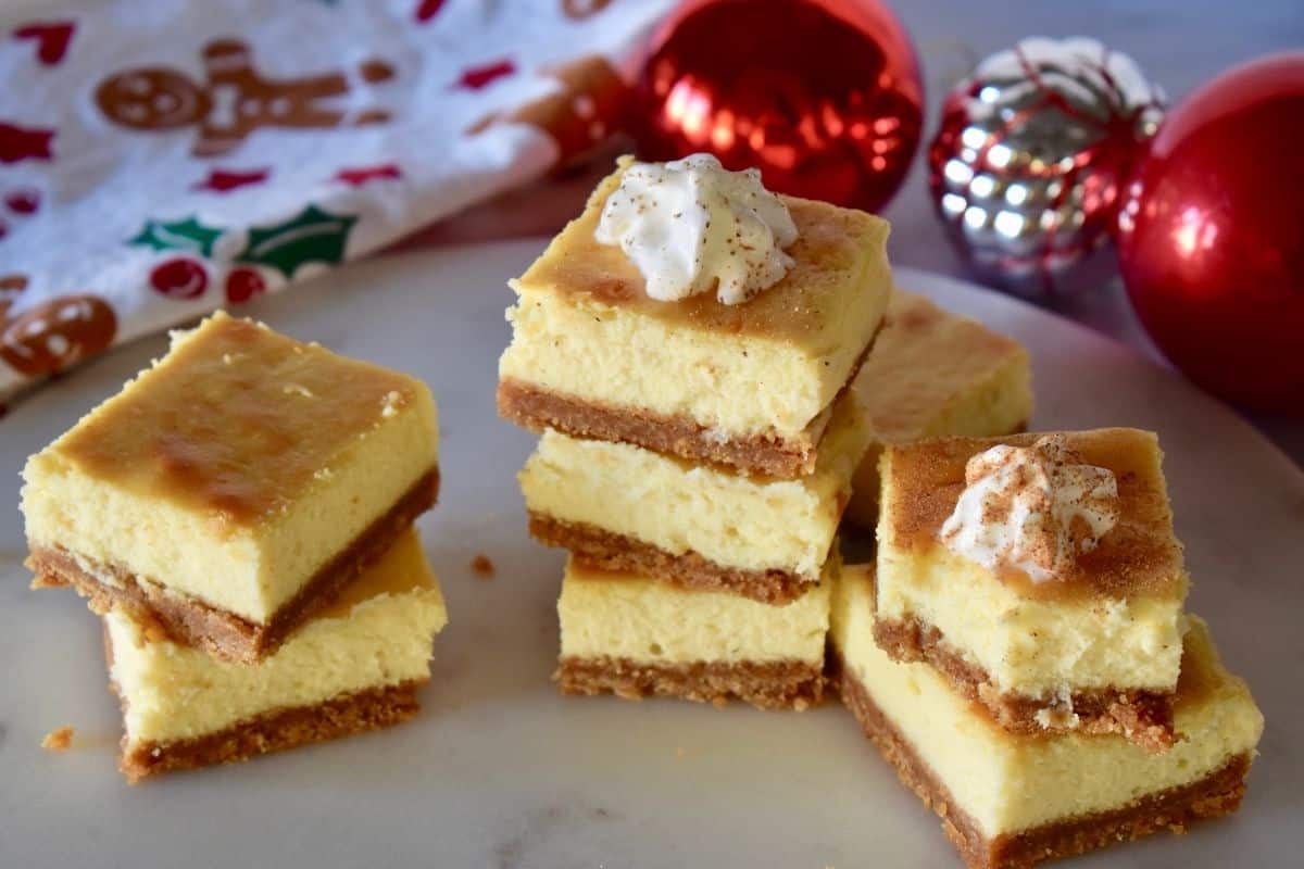 stacks of cheesecake bars on a white serving platter with red ornaments in the background. 