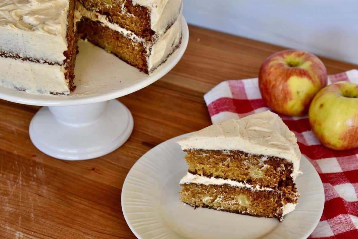 slice of apple spice cake on a white plate with pedestal of the cake next to it. 