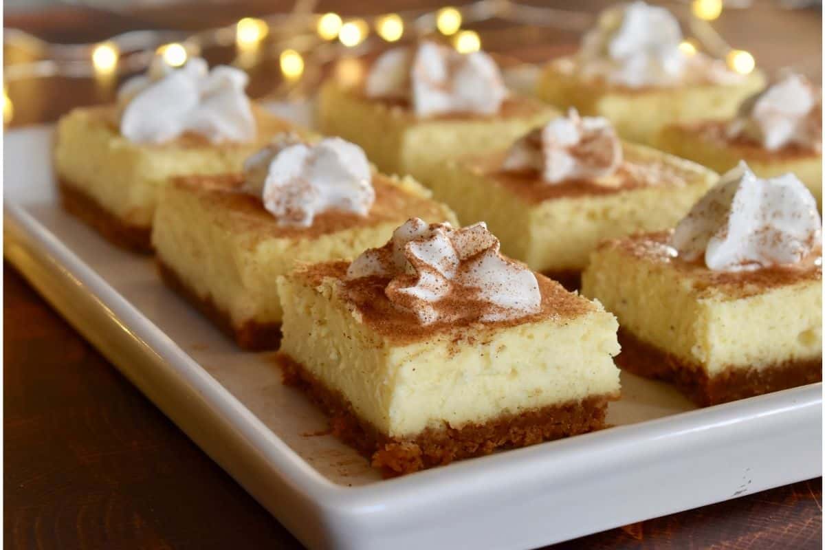 Prepared cheesecake bars on cut on a white serving platter with dollops of whipped cream on top. 