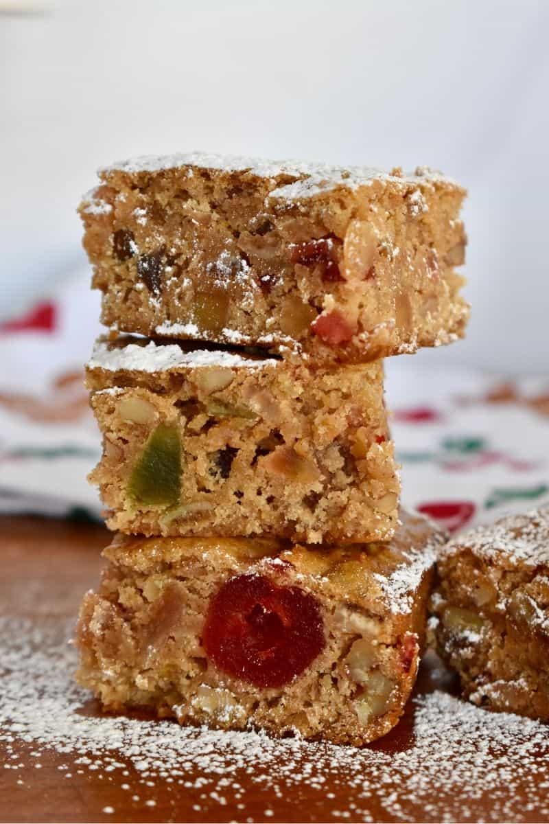 Fruitcake cookie bars stacked high on a cutting board with powdered sugar sprinkled on them. 