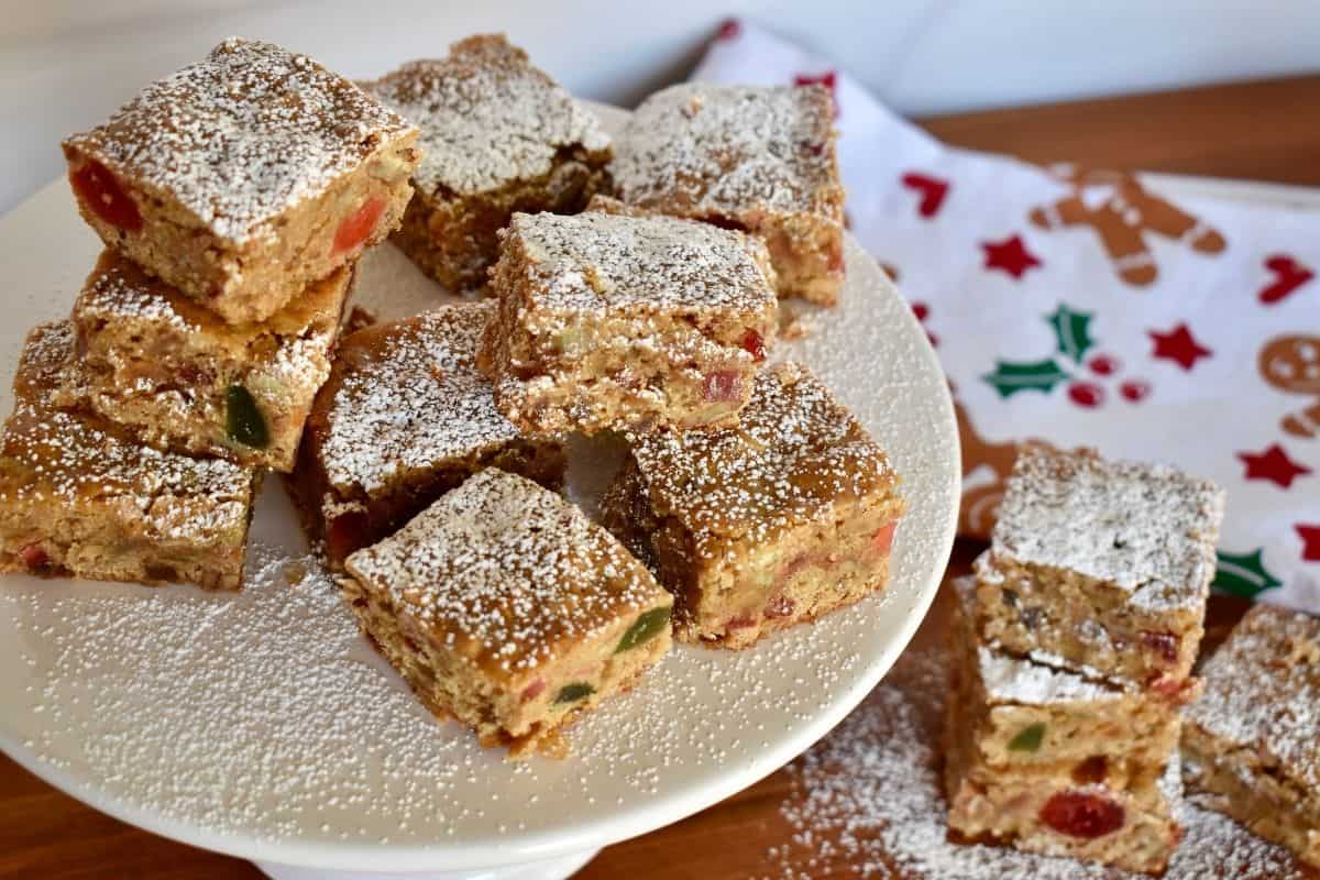 Fruitcake cookie bars on a white pedestal covered in powdered sugar. 