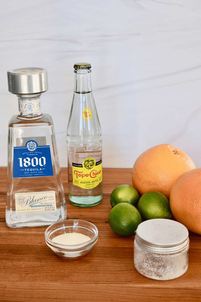 vertical photo of ingredients needed to make cocktail including tequila, limes, salt, and mineral water. 