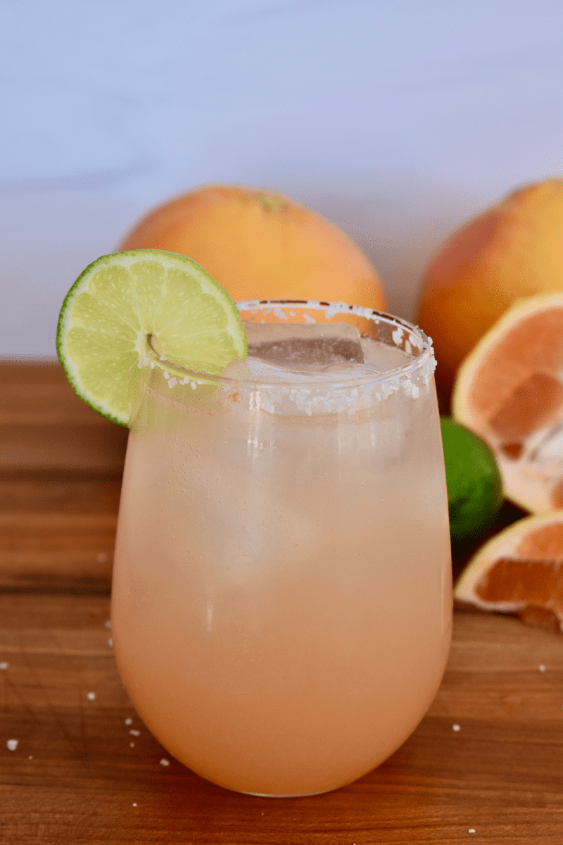 Grapefruit Paloma in a glass with a slice of lime on the rim and grapefruits in the background. 