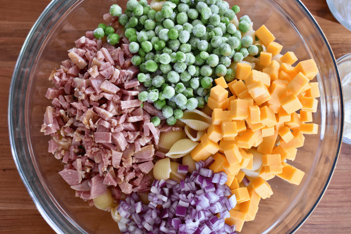 overhead photo of ingredients for recipe in a bowl including noodles, peas, cheddar, and red onion. 