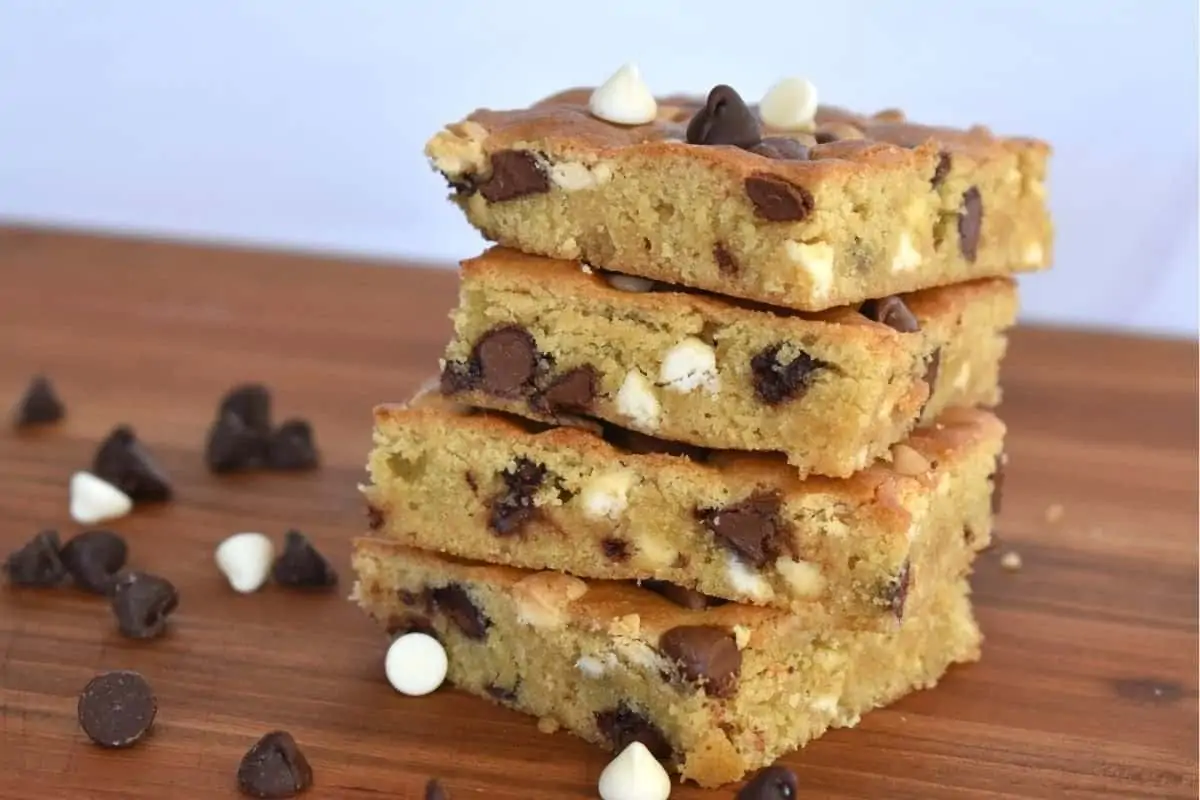 Sheet Pan Cookie Bars stacked on each other on a wood cutting board with white chocolate and milk chocolate chips. 