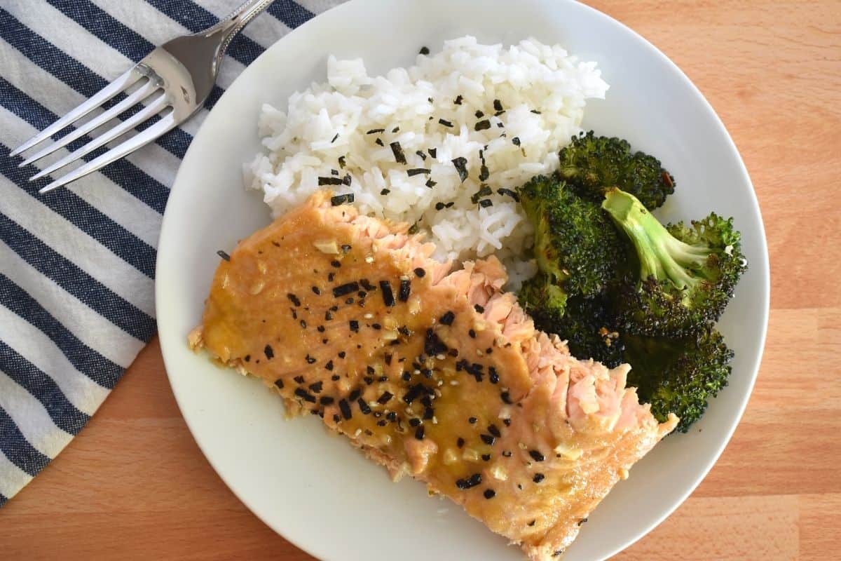 Asian Salmon Marinade fish on a plate with rice and broccoli with a striped napkin and fork in the background. 