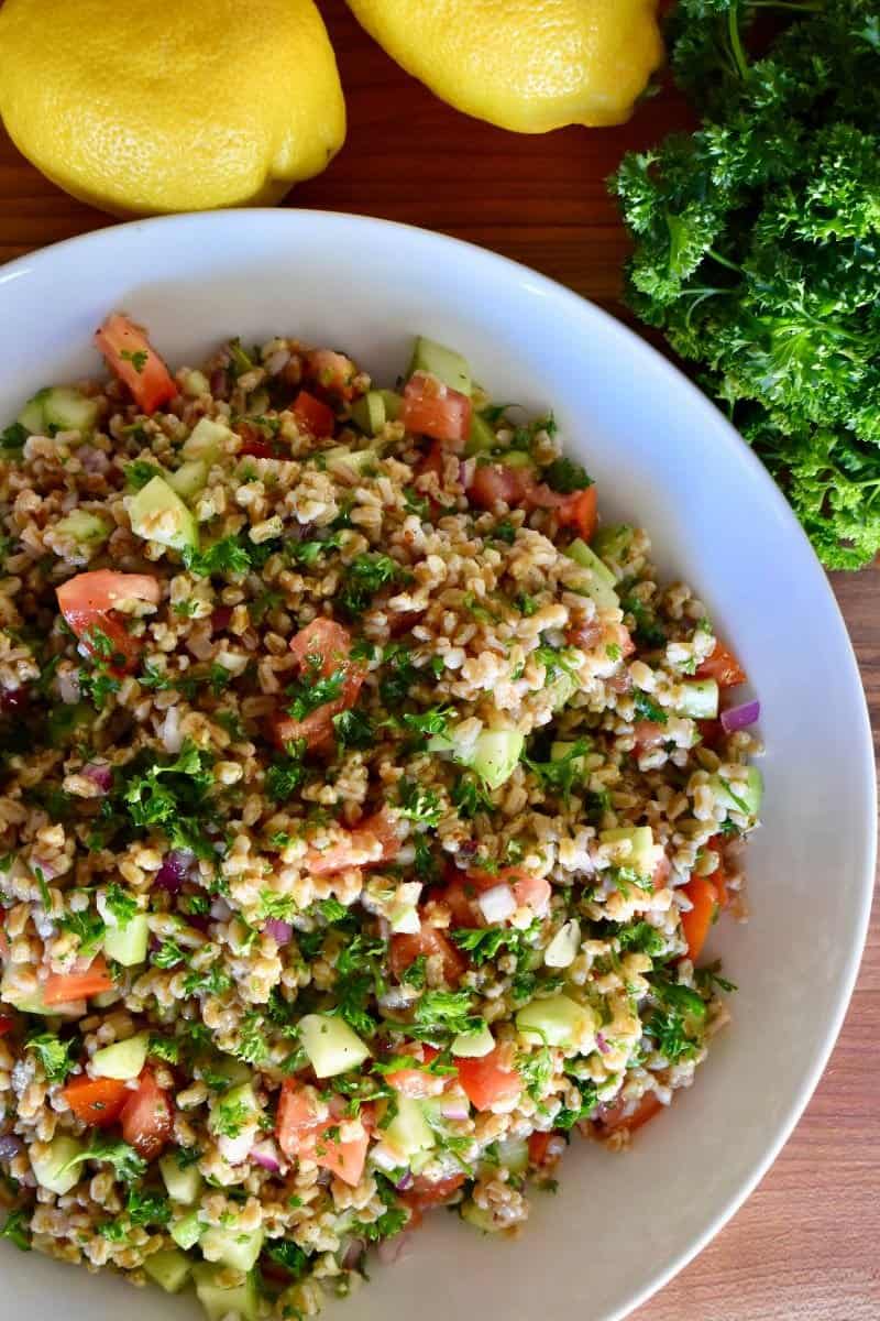 overhead photo of farro tabbouleh salad in a white bowl with lemons and parsley next to it. 
