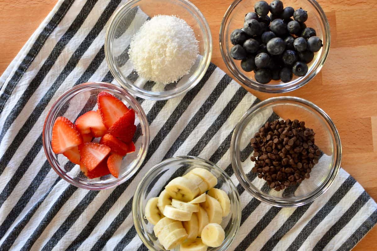 overhead photo of toppings for the recipe including strawberries, blueberries, mini chocolate chips, bananas, and coconut.  