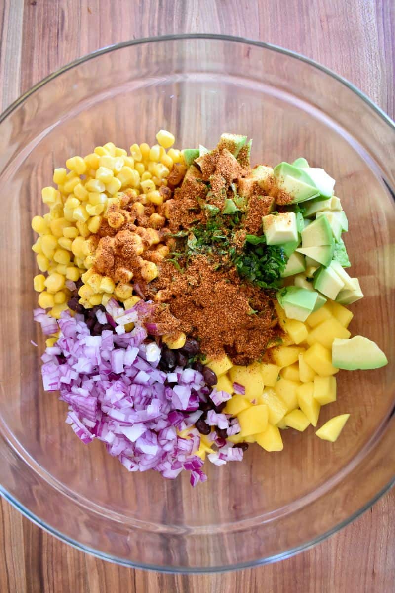 overhead photo of ingredients for the recipe in a glass bowl including cilantro, avocado, corn, and red onion. 