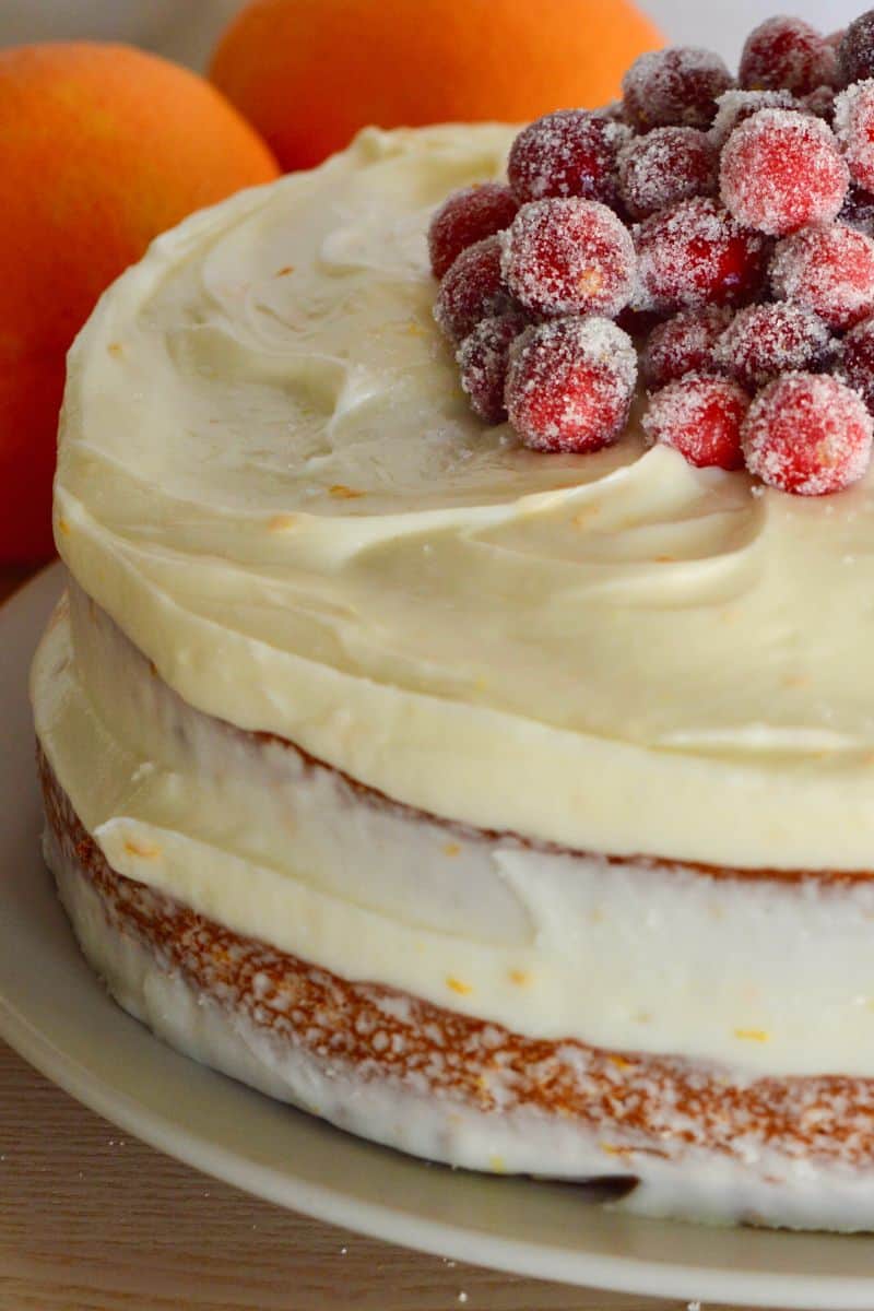 Cranberry Orange Cake with cream cheese frosting and sugared cranberries with oranges behind it. 