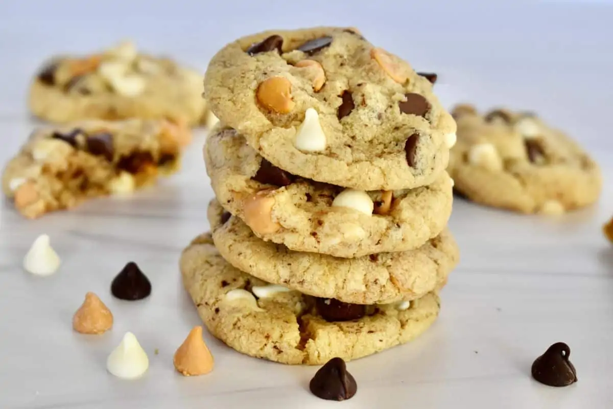 Triple Chocolate Chip Cookies stacked on each other on a white surface with chips sprinkled around them. 