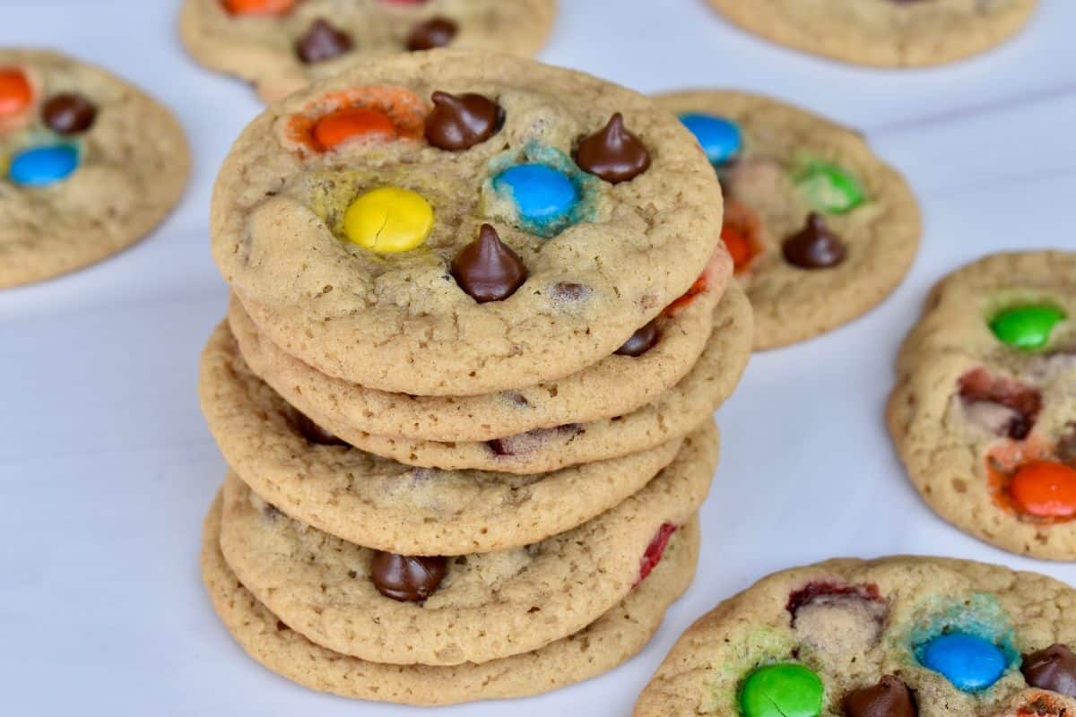 A stack of M&M Chocolate Chip Cookies on a white countertop with more cookies in the background. 