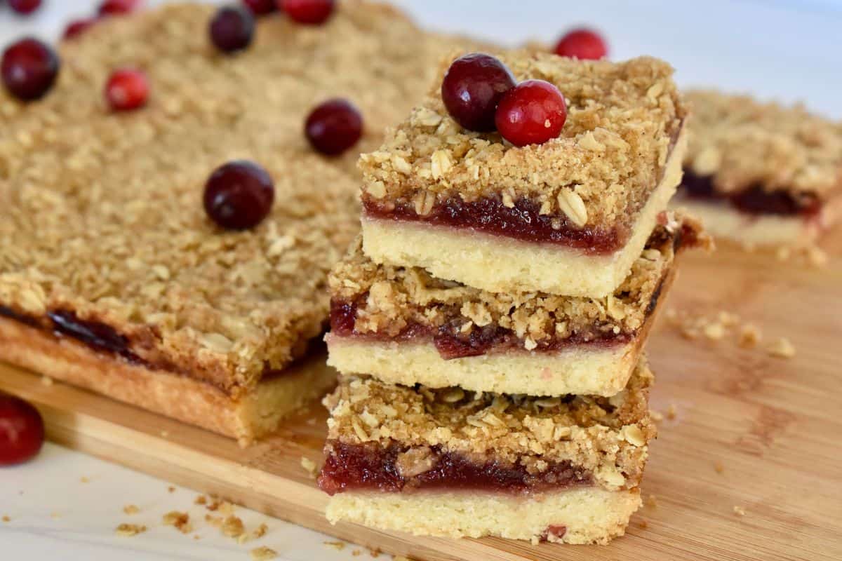 Cranberry Shortbread Bars stacked on each other on a wood cutting board with cranberries on it. 