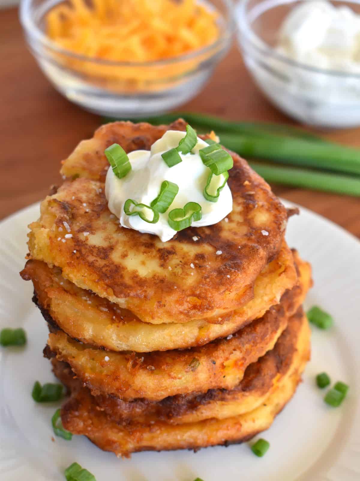 Mashed Potato cakes on a white plate with cheddar cheese and sour cream in the background. 