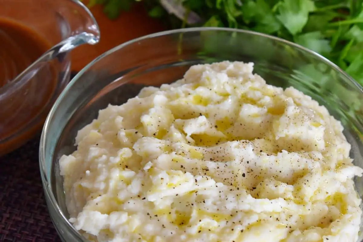 Ricotta Mashed Potatoes in a glass mixing bowl with parsley and gravy in the background. 