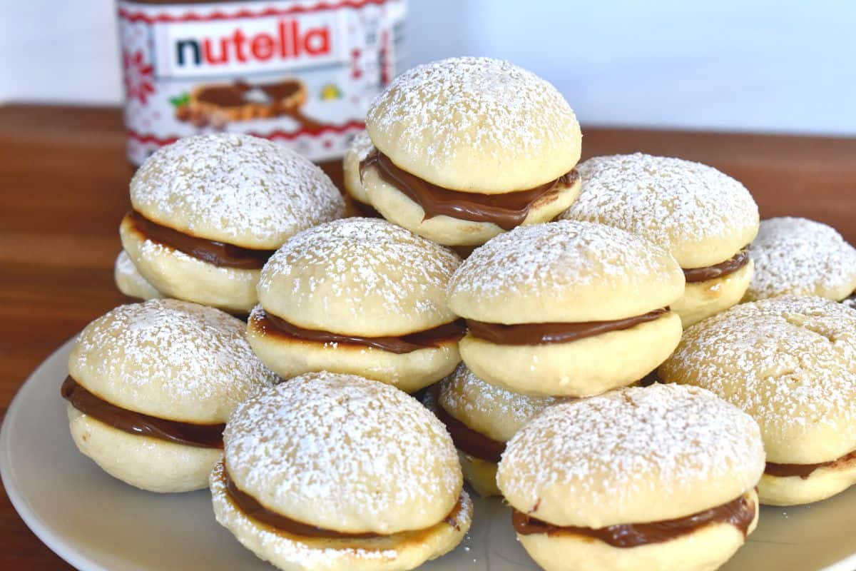 Nutella Sandwich Cookies stacked on each other on a white plate with a jar of Nutella in the background. 
