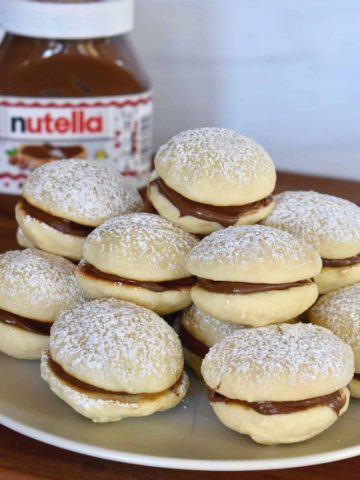 Nutella Sandwich Cookies with Nutella filling piled on a plate with a jar of Nutella behind it.