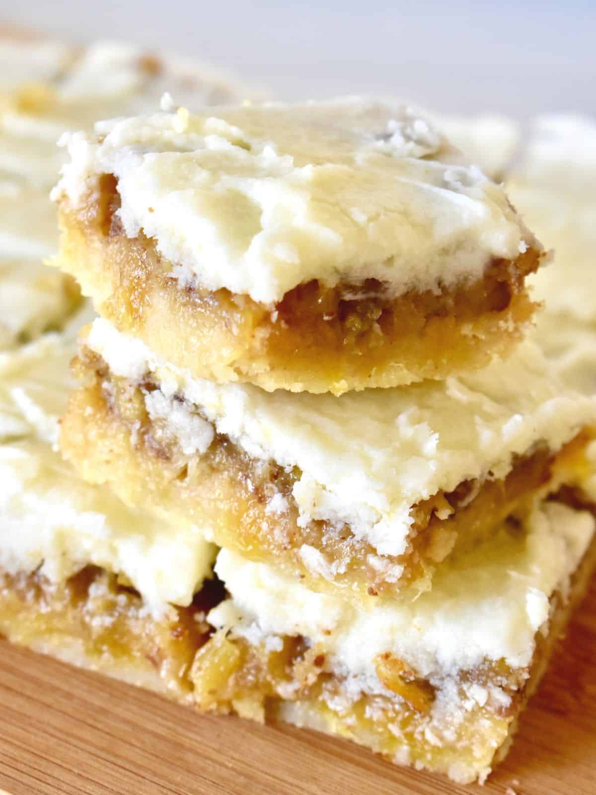 Pineapple bars stacked on each other with frosting on top and more squares in the background. 