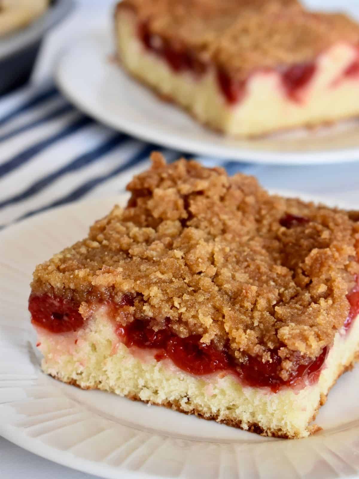 Cherry Coffee Cake slice on a white plate with a slice in the background and striped napkin. 