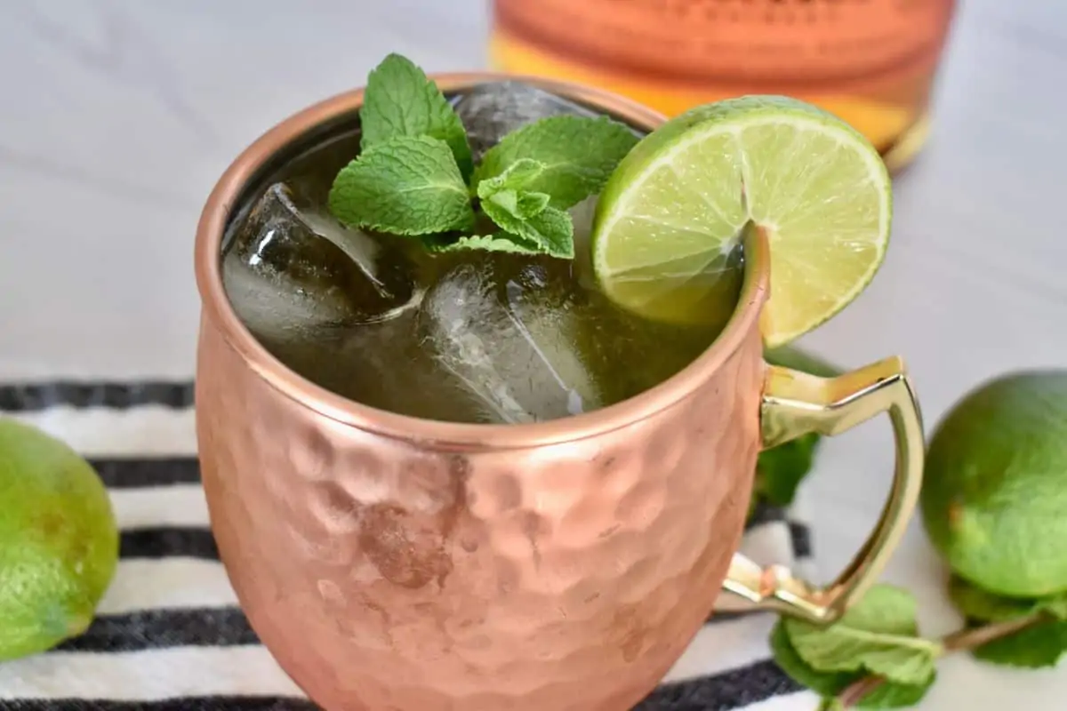 Kentucky Mule in a copper mug garnished with mint and lime and a bottle of bourbon in the background. 