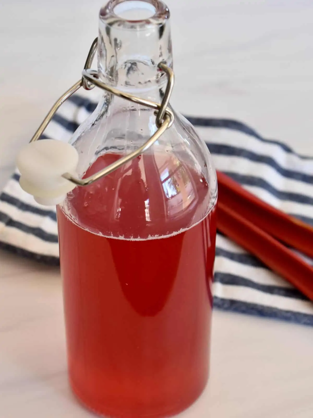 Rhubarb Simple Syrup in a glass container with a stalk of rhubarb and striped napkin behind it. 
