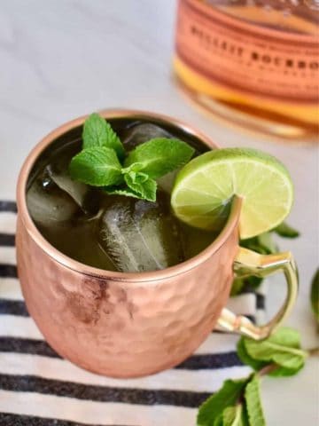 Kentucky Mule in a cooper mug full of ice garnished with mint and lime with a bottle of bourbon in the background.