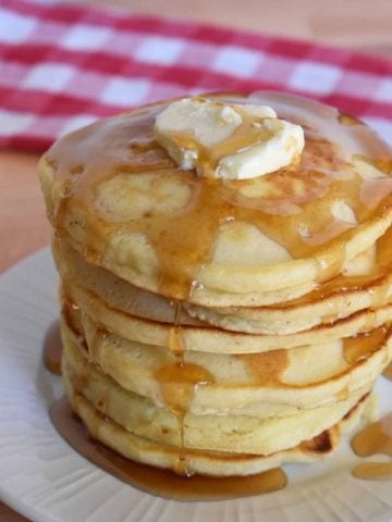 stack of pancakes piled on a white plate with butter and syrup on top.